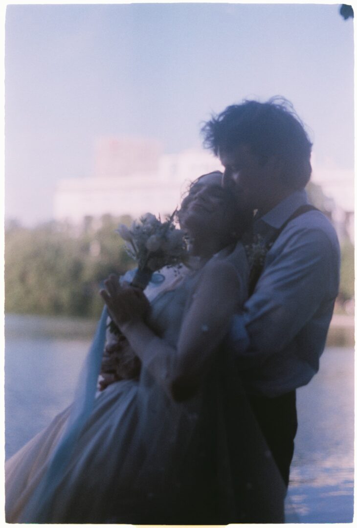 Bride and groom embracing closely by a lake at sunset, softly silhouetted against the water and evening sky