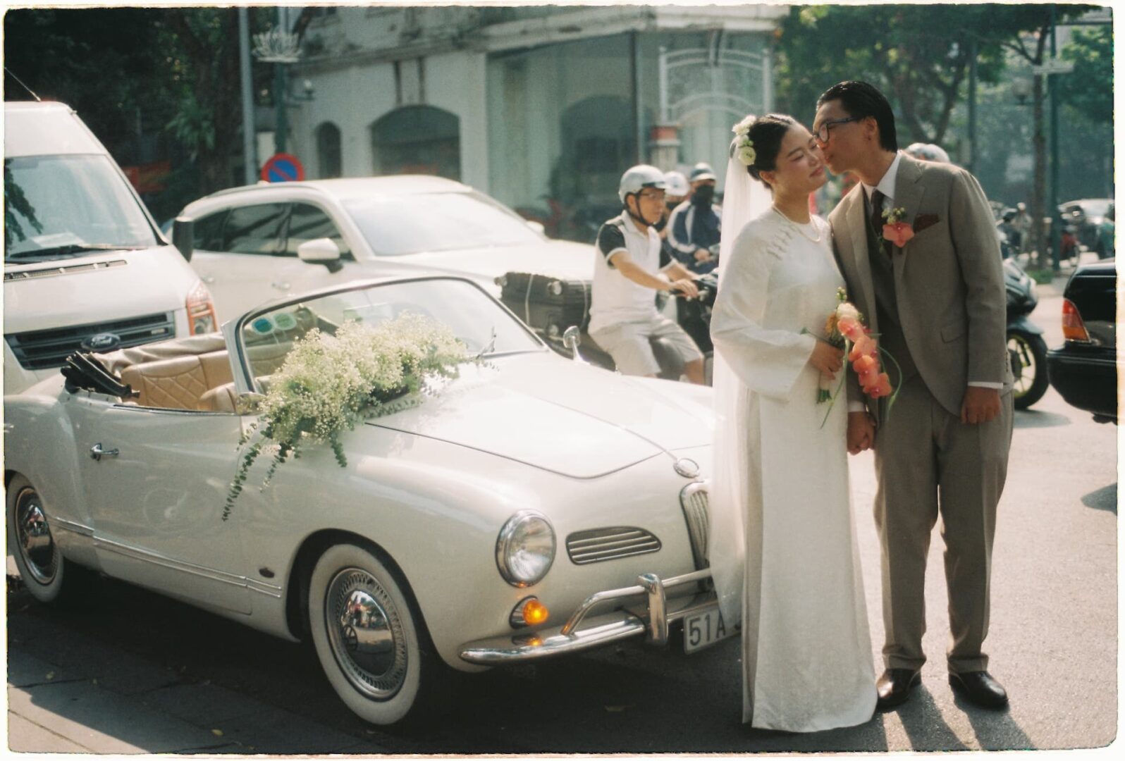 Bride and groom standing together beside a white vintage convertible decorated with flowers on a city street on their wedding day