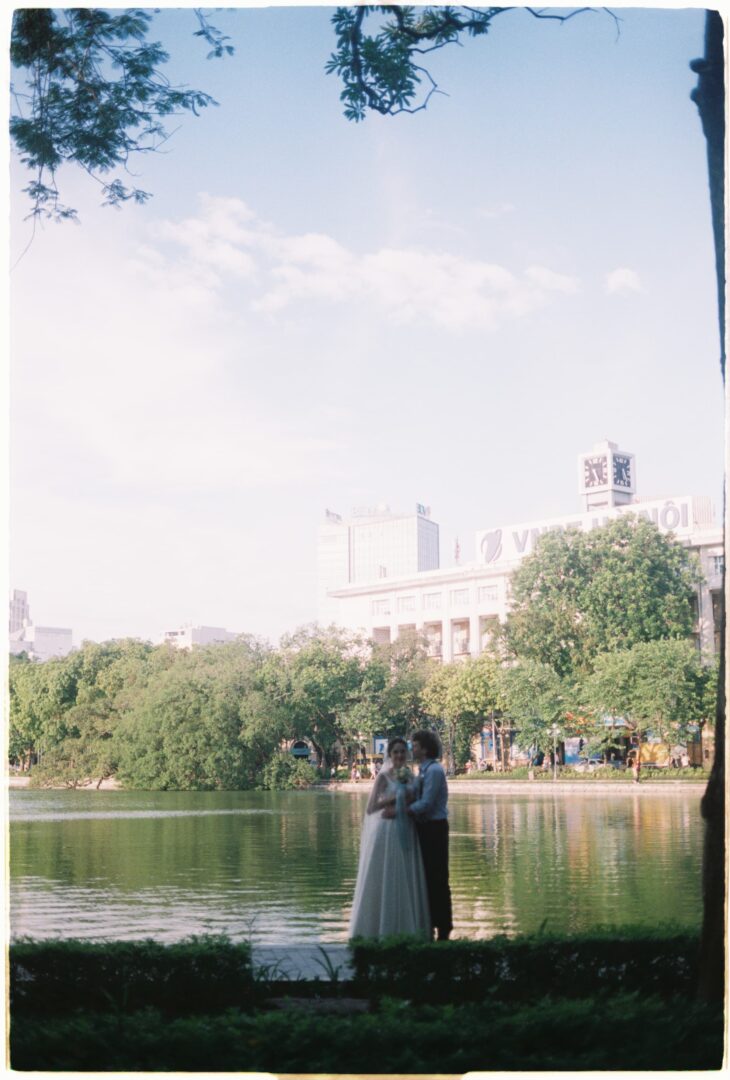 Bride and groom standing together by a lakeside in a city park, surrounded by trees and reflected water