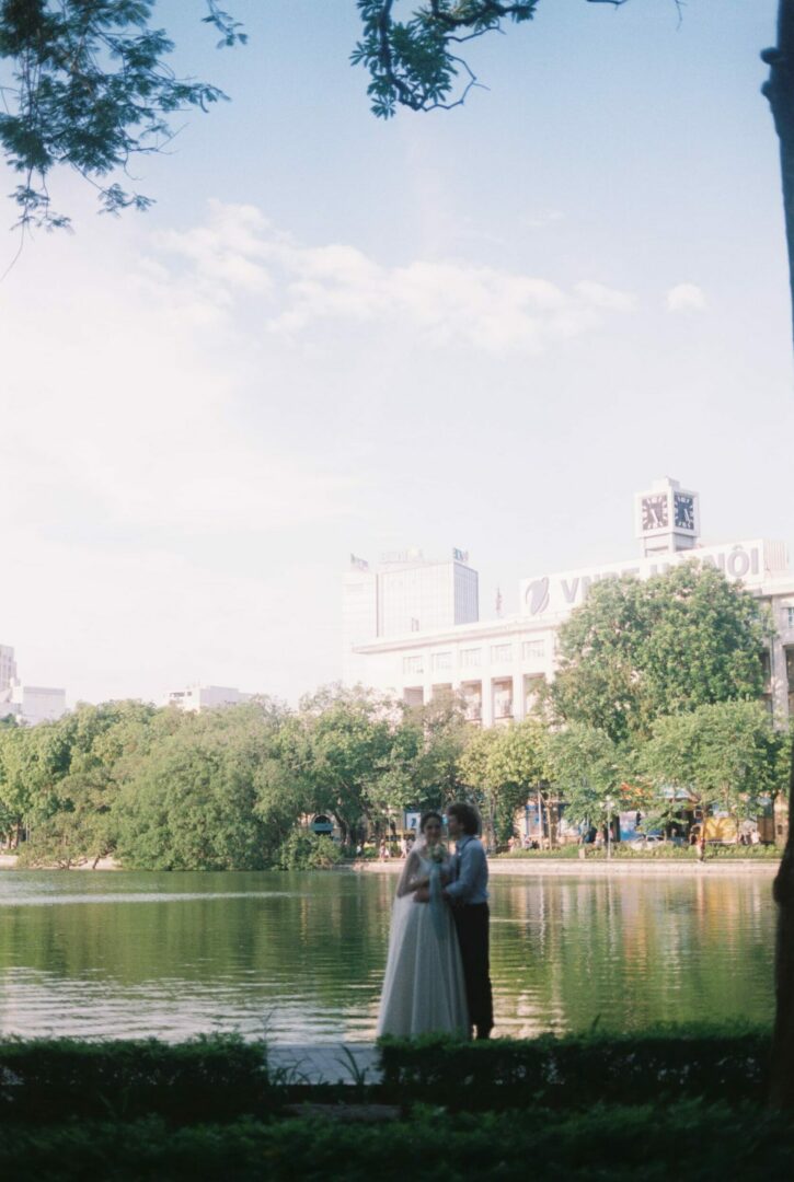 Bride and groom standing together by a lakeside in a city park, surrounded by trees and reflected water