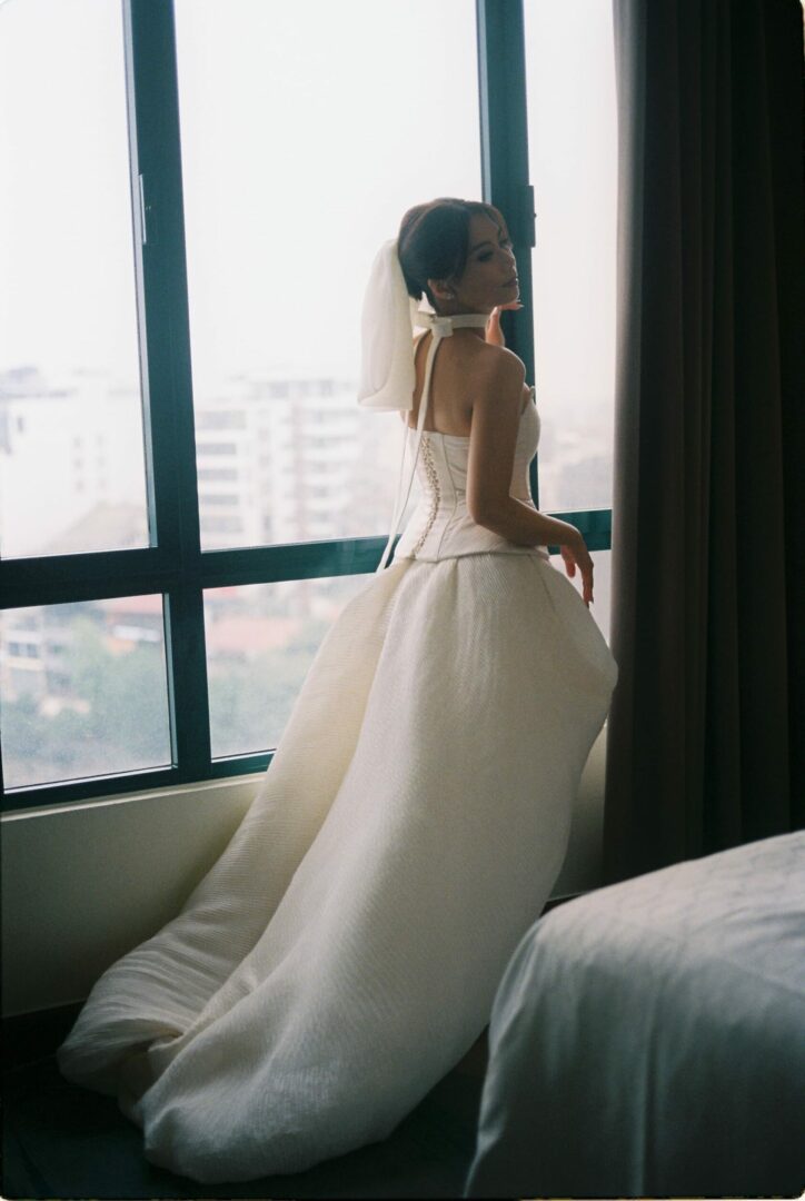 Bride in a white wedding dress standing by a large window in a hotel room, softly lit by natural daylight before the ceremony