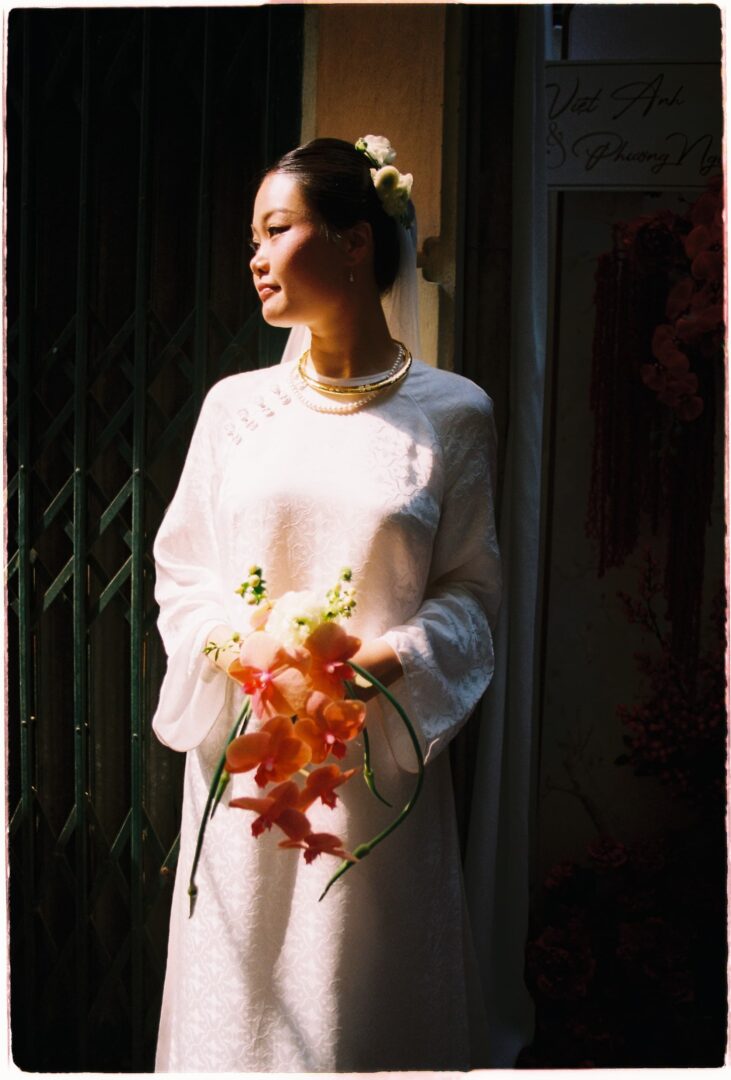 Bride wearing a traditional white wedding dress, holding an orchid bouquet and standing in soft natural light near an entrance