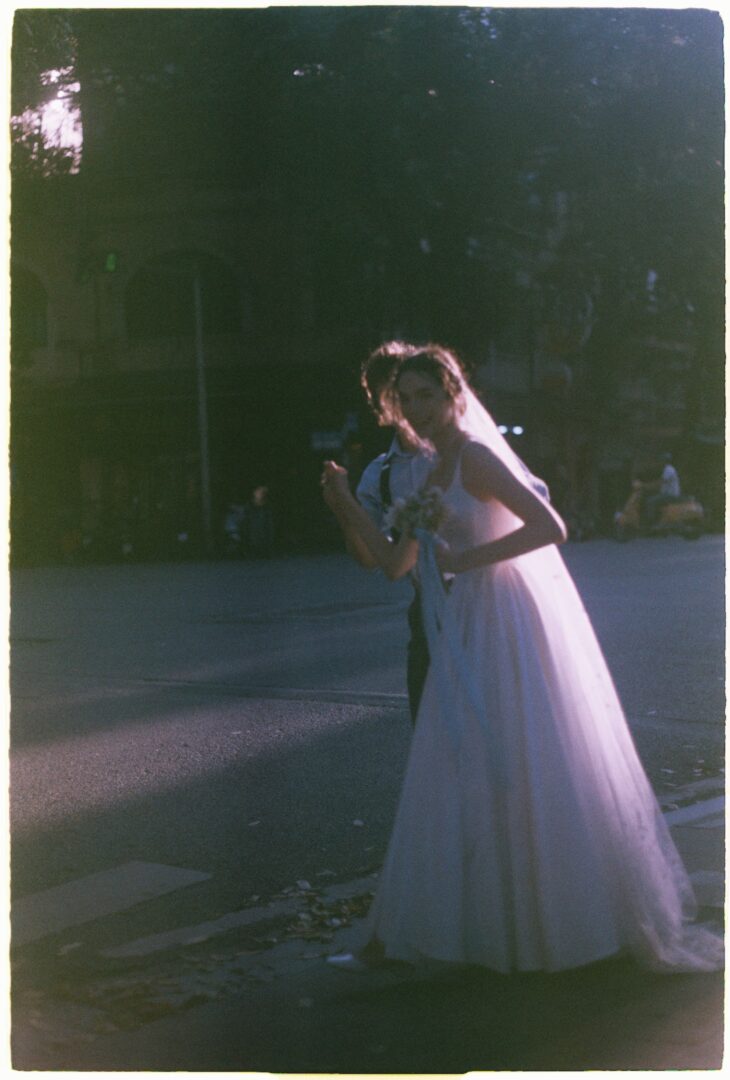 Bride in a flowing white wedding dress walking outdoors at dusk, softly backlit by natural light