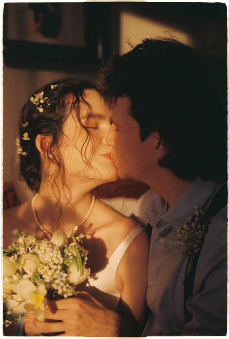 Bride and groom sharing an intimate kiss indoors, bathed in warm golden light, holding a bouquet of white flowers