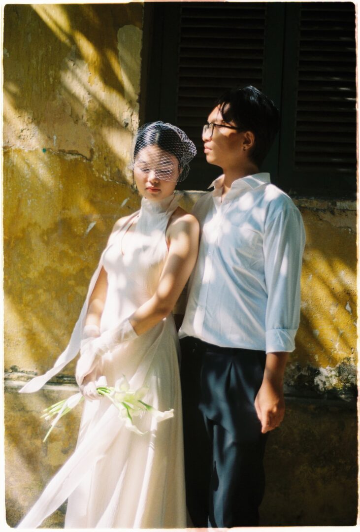 Bride in a minimalist white dress with a veil standing beside the groom against a textured wall, softly lit by dappled sunlight
