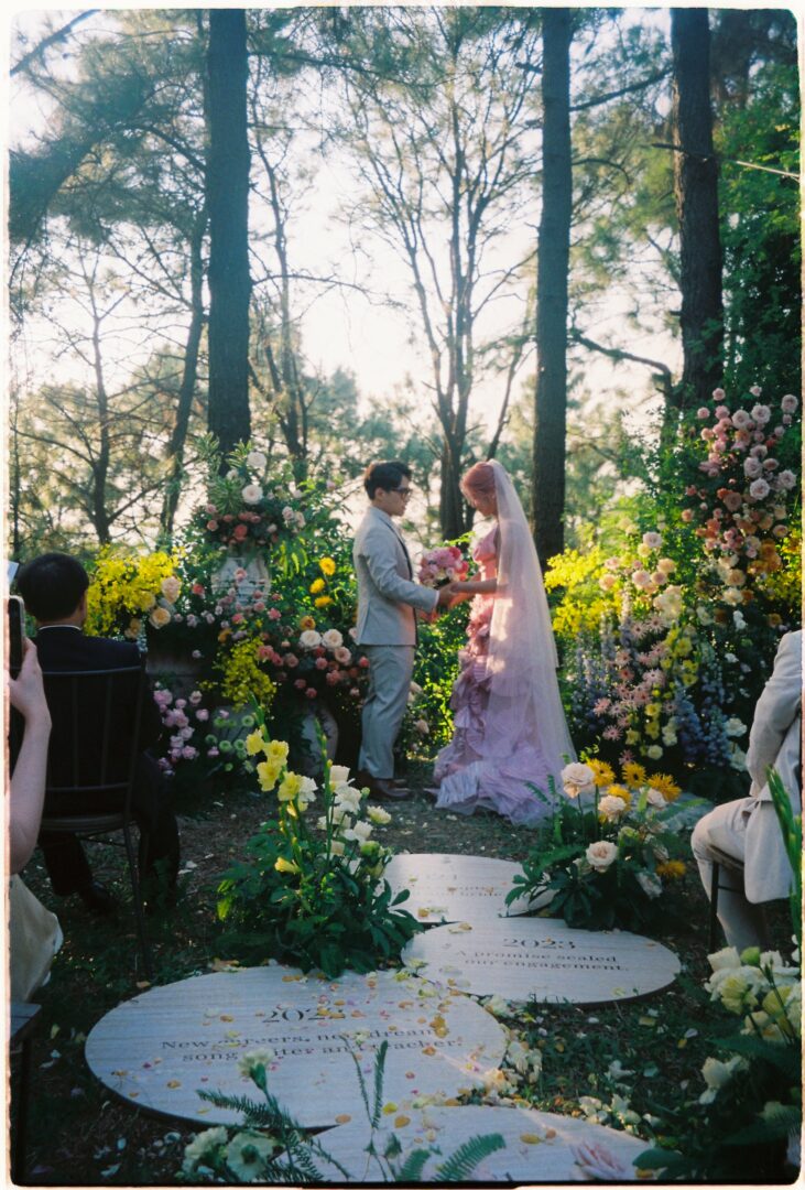 Bride and groom exchanging vows during an outdoor wedding ceremony in a forest setting, surrounded by colorful floral arrangements and guests