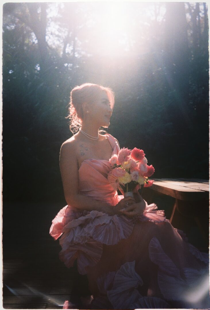 Bride in a pink wedding dress holding a bouquet, seated outdoors in a forest setting and softly backlit by warm sunset light